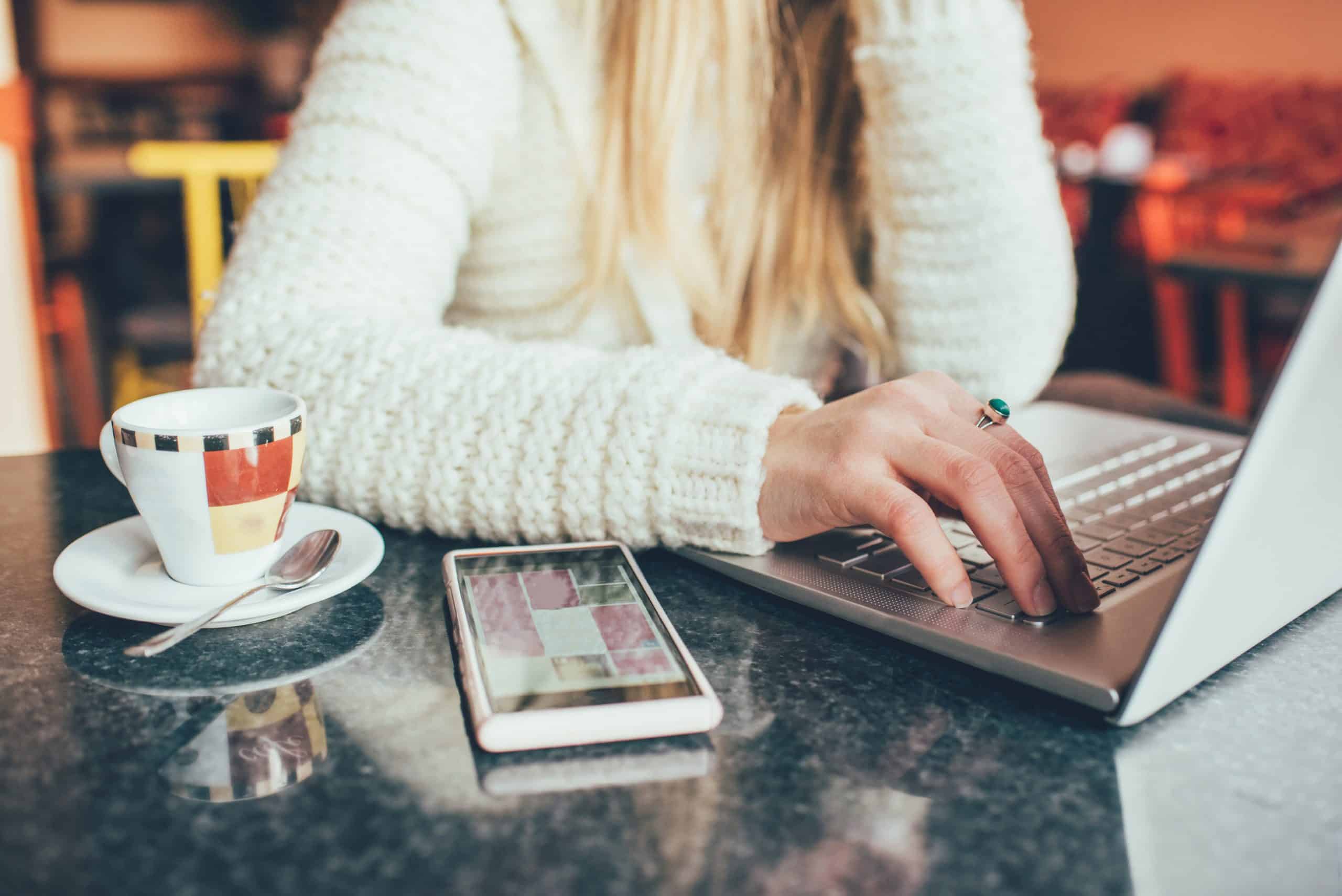 Close up on the hand of a young woman sitting on a bar having a coffee, using smart phone and tapping pc keyboard - leisure, technology, social network concept
