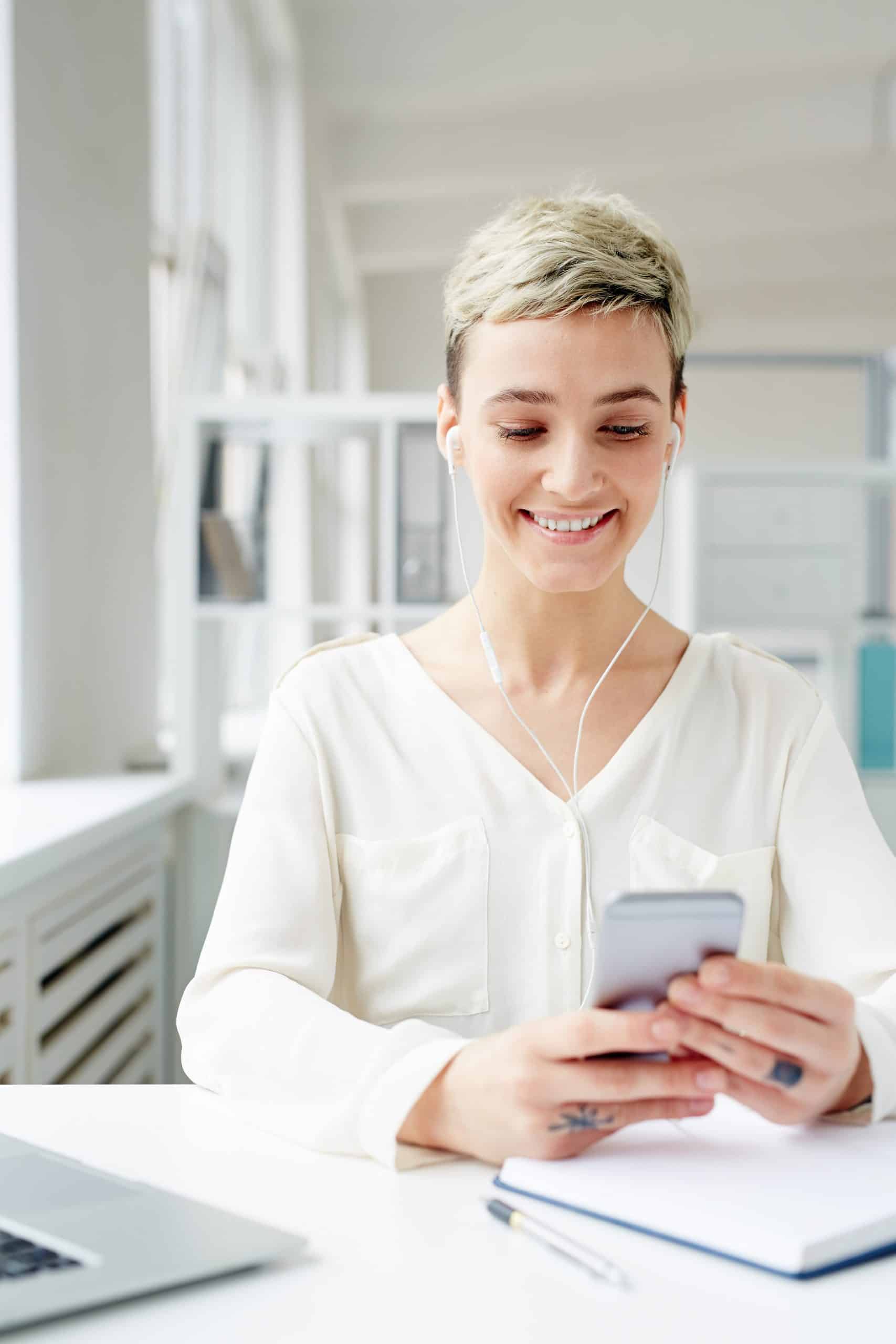 Positive young businesswoman choosing her favorite tracks from playlist in smartphone by workplace